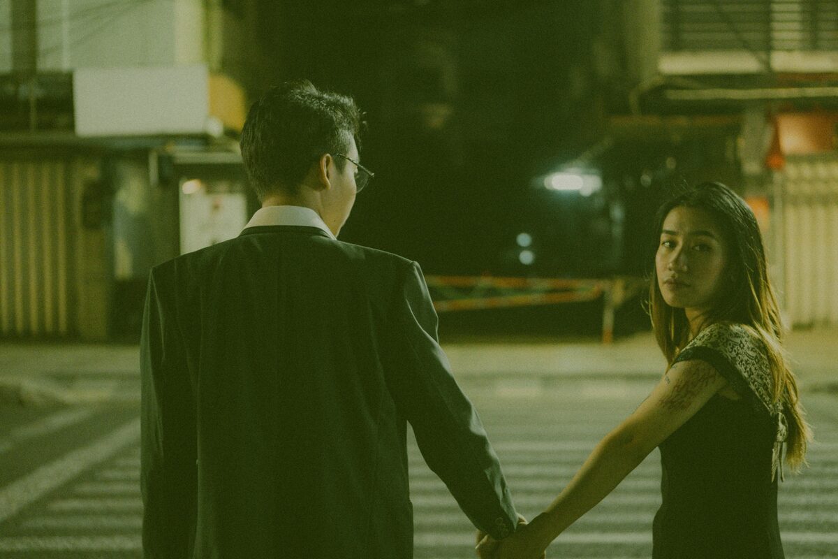 A couple holding hands while crossing a city street at night under urban lights.