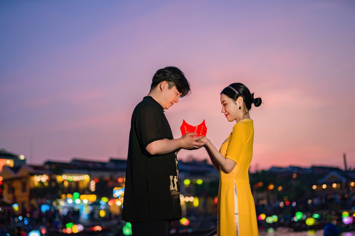 Romantic couple exchanging a red lantern at sunset with a vibrant city skyline in the background.