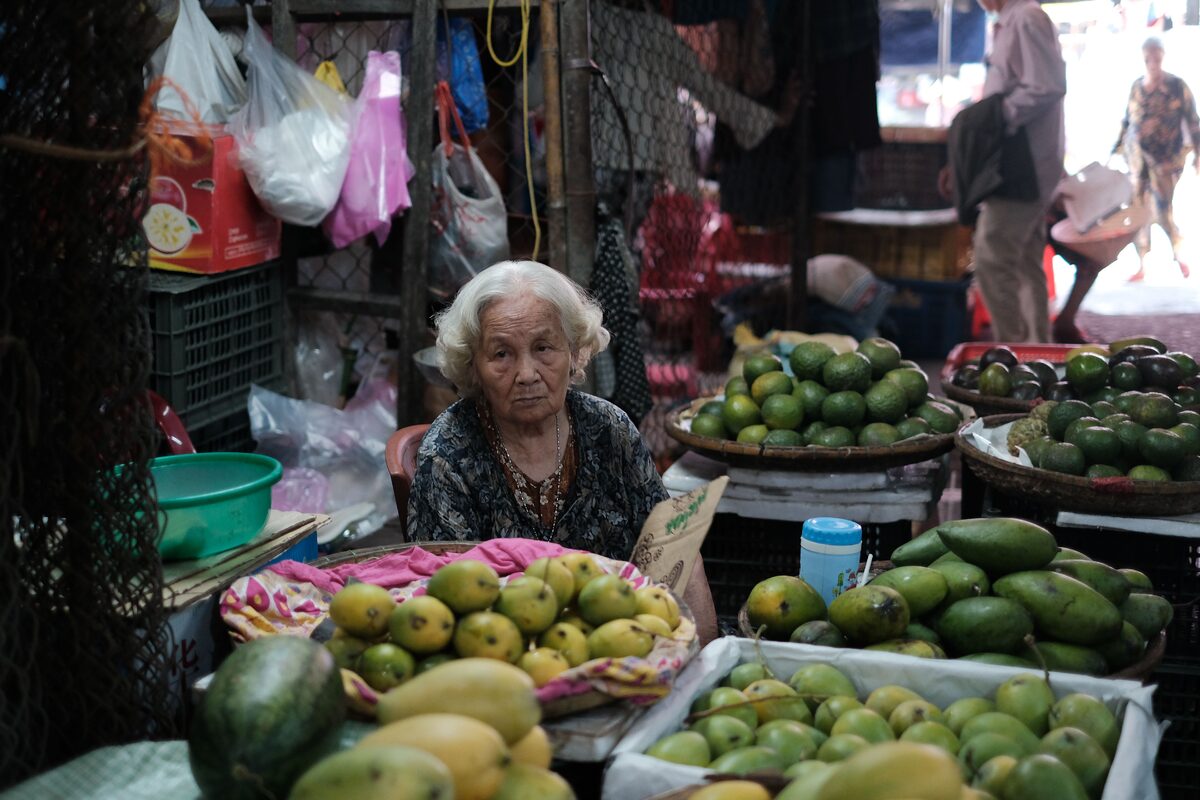 An elderly woman selling fresh mangoes and avocados at an indoor market fruit stall.