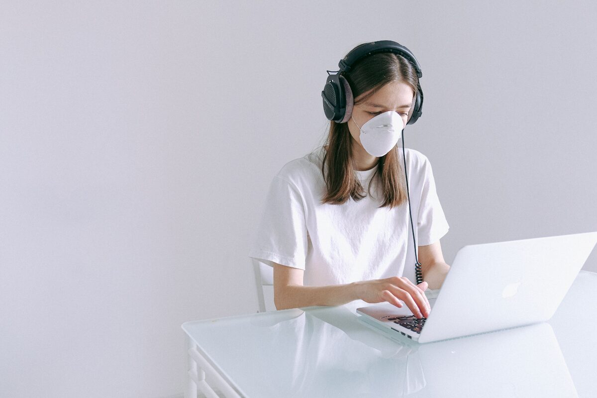 Woman wearing a face mask and headphones working on a laptop at home, representing remote work during the pandemic.