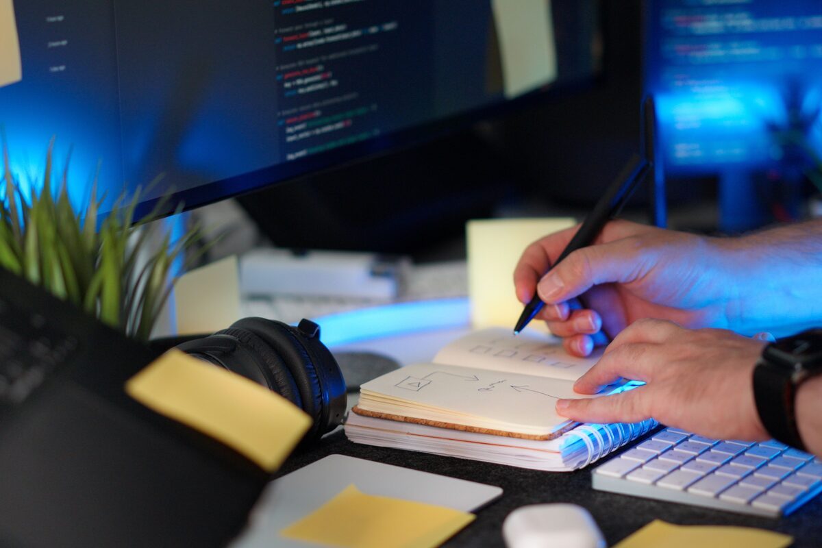 A person planning a software project with handwritten notes and code on a monitor in a modern workspace.