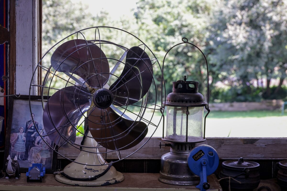 A vintage fan and lantern on a wooden shelf with a picturesque outdoor view.