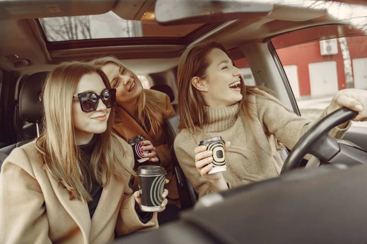 Happy young female friends in stylish casual clothes driving around city streets while drinking takeaway coffee and having fun