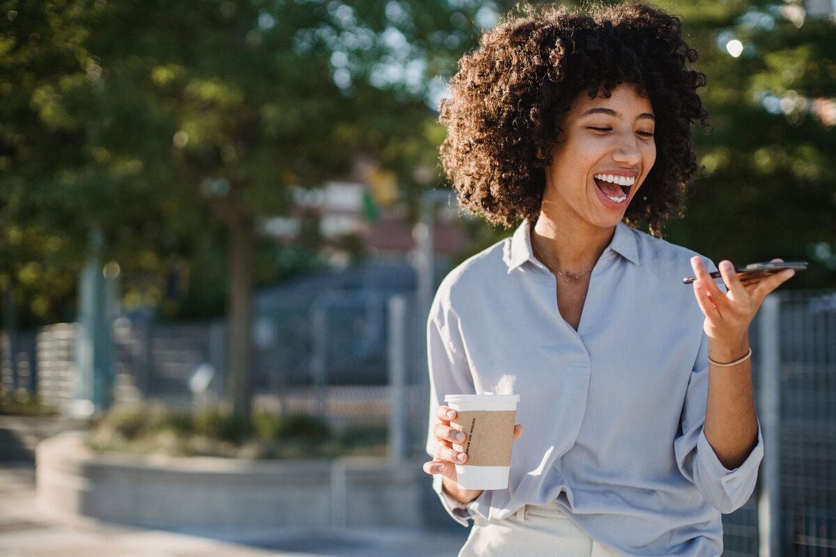 Smiling woman enjoying a sunny day while using her smartphone and drinking coffee outdoors.