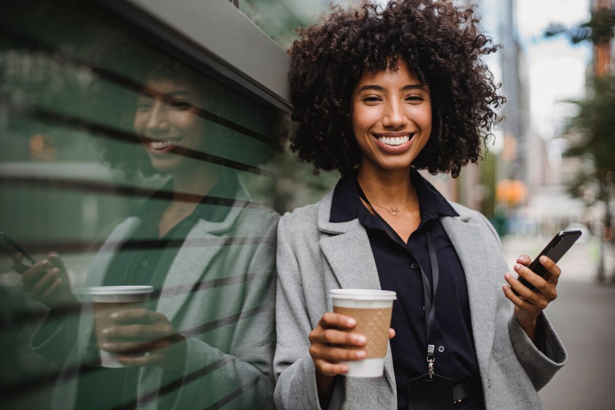 Young woman holding coffee and smartphone, smiling on a city street.