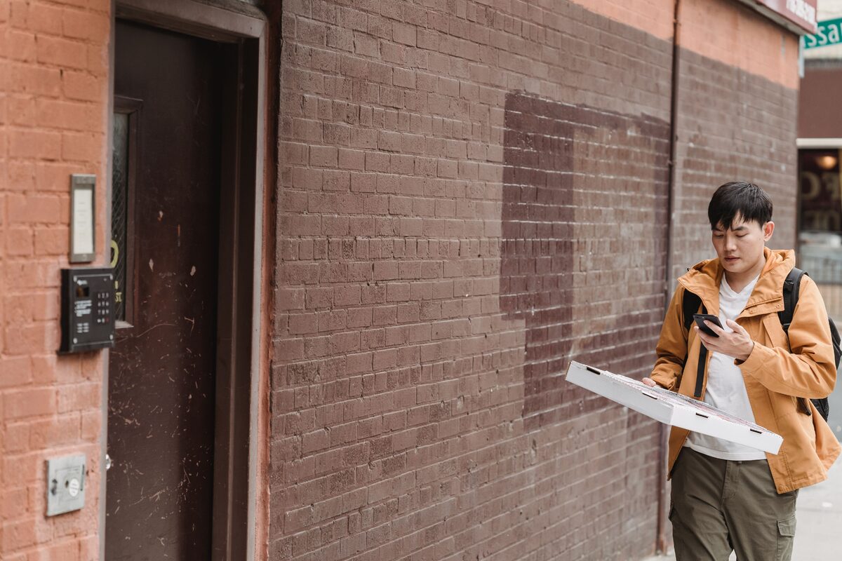 Asian delivery man with pizza box checking smartphone near urban doorstep.