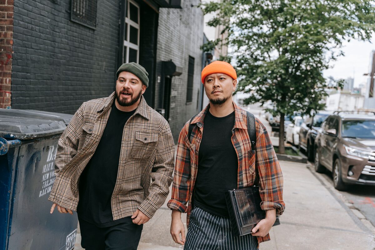 Two men in plaid shirts walk down a city sidewalk, engaging in conversation.