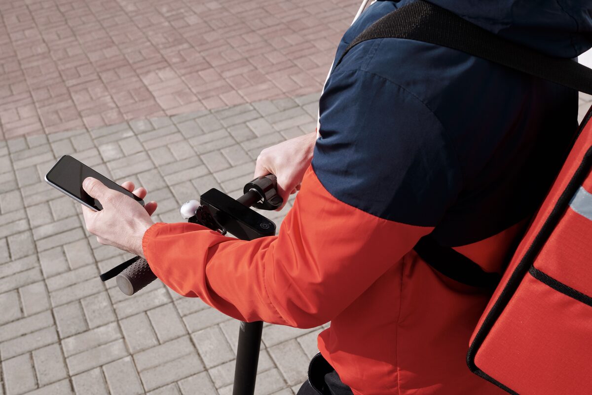 A delivery person riding an electric scooter using a smartphone for navigation outdoors.