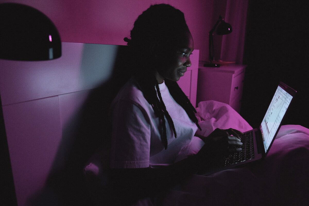 An adult woman using a laptop in bed under purple lighting, representing modern remote work at home.