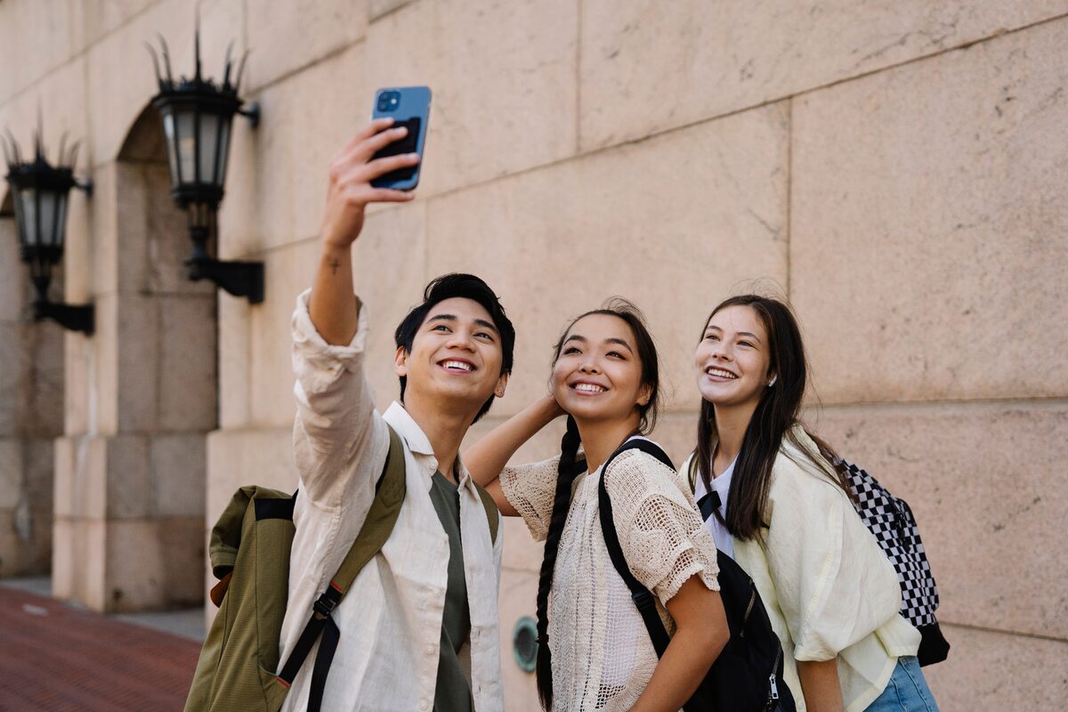 Three young friends smiling and taking a selfie outdoors with a smartphone.