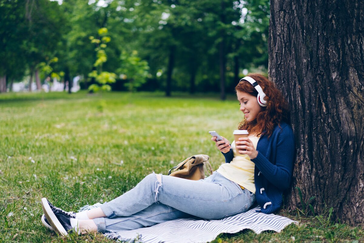 Young woman enjoying a sunny day in the park with headphones, coffee, and smartphone.