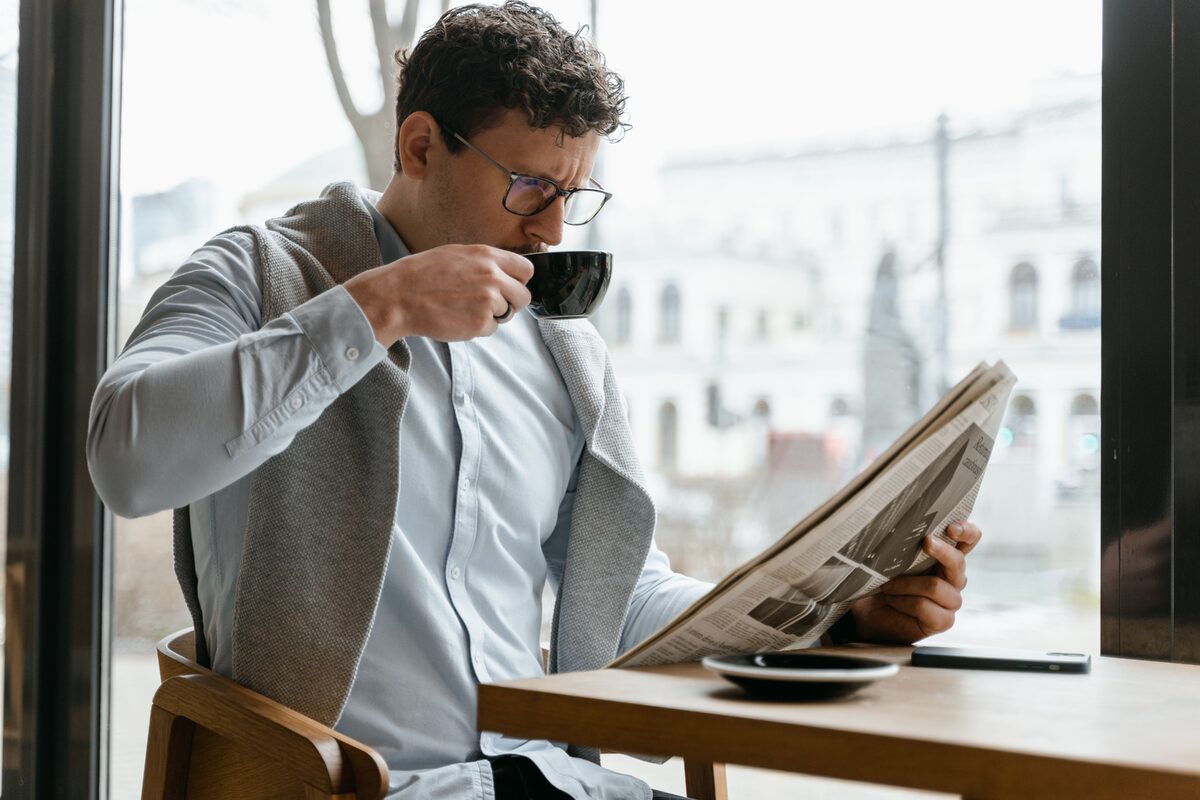 Adult man wearing eyeglasses sipping coffee and reading a newspaper in a cozy cafe setting.