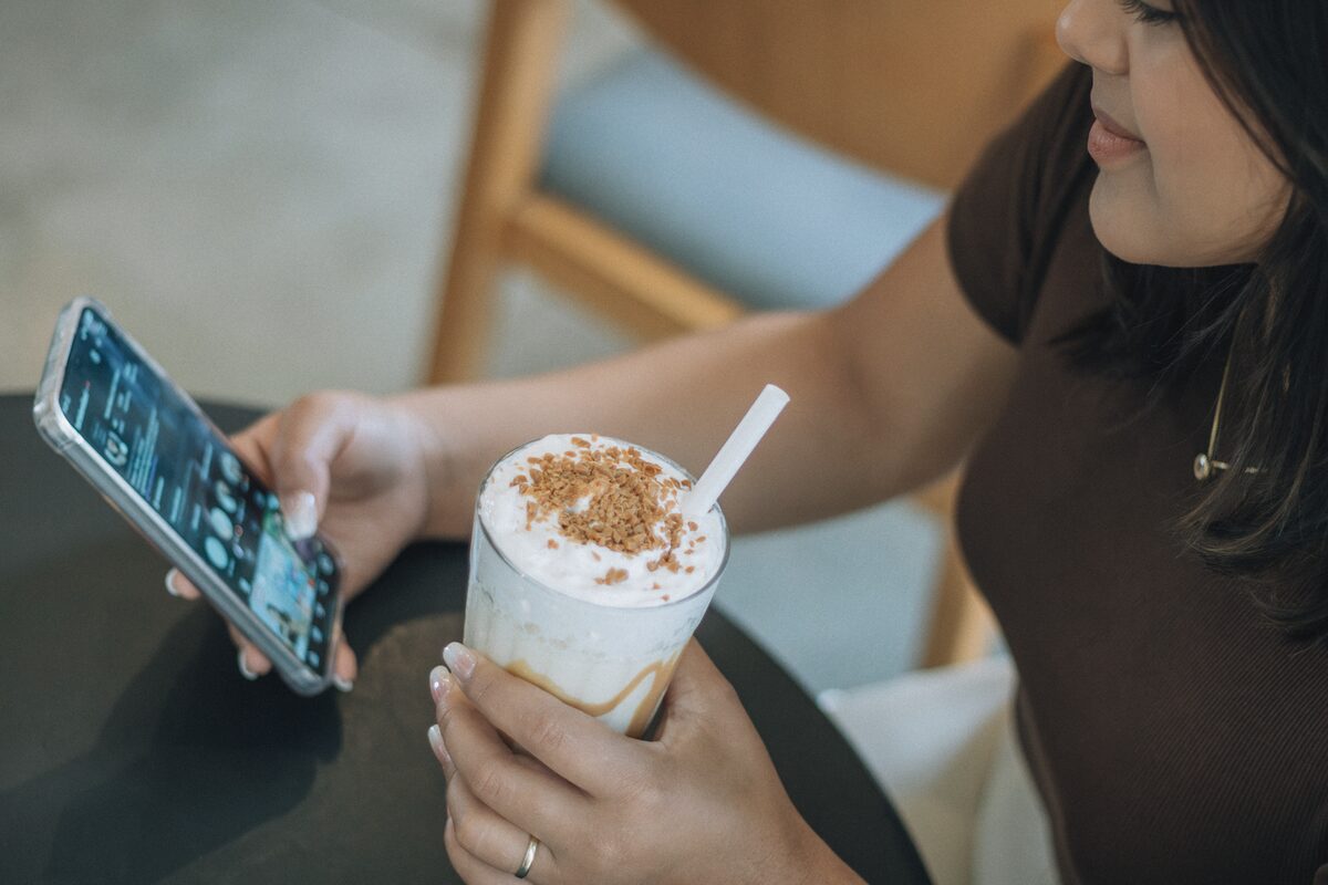 A woman is sipping iced coffee and browsing her smartphone at a cafe.
