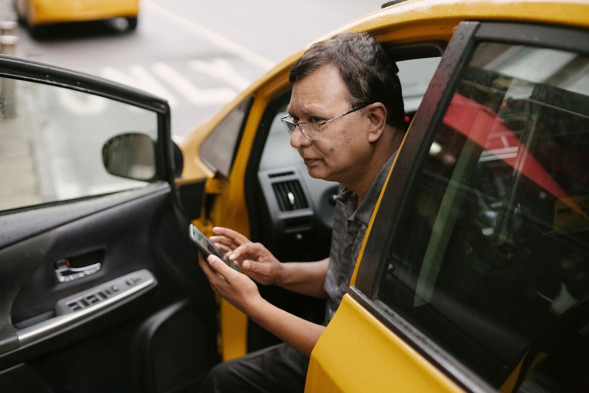 Crop Asian man sitting in parked yellow taxi car with open door and using smartphone while looking away
