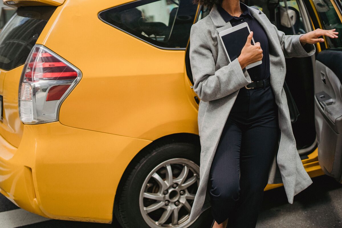 Crop unrecognizable female in formal clothes holding tablet while getting out of yellow taxi