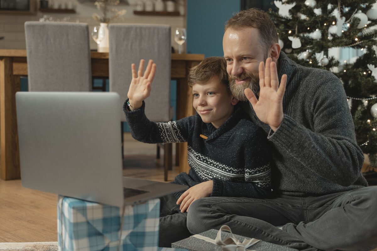Father and son waving during a video call at home with Christmas decorations and gifts.