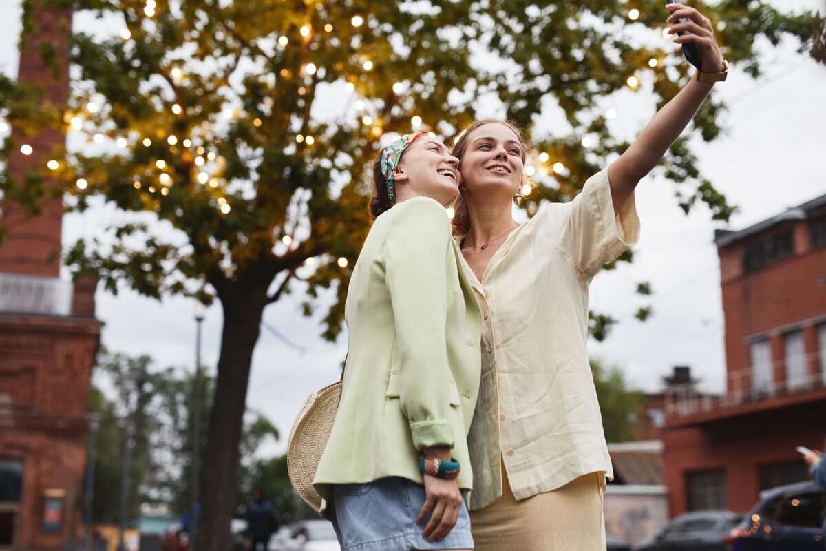 Two young women taking a selfie outdoors with a festive tree background in an urban setting.