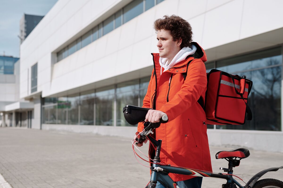A bicycle delivery worker in a red jacket stands with a bike outdoors, ready for deliveries.